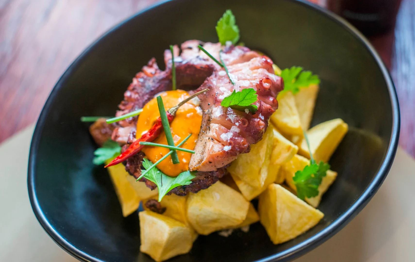 Presentación de un plato de pulpo seco ahumado encima de unas patas bravas, acompañado de un poco de salsa, guindilla y perejil fresco.