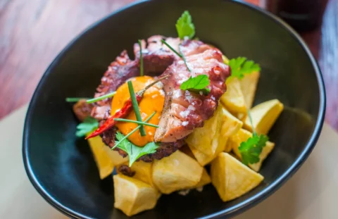 Presentación de un plato de pulpo seco ahumado encima de unas patas bravas, acompañado de un poco de salsa, guindilla y perejil fresco.