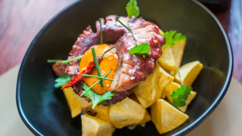 Presentación de un plato de pulpo seco ahumado encima de unas patas bravas, acompañado de un poco de salsa, guindilla y perejil fresco.